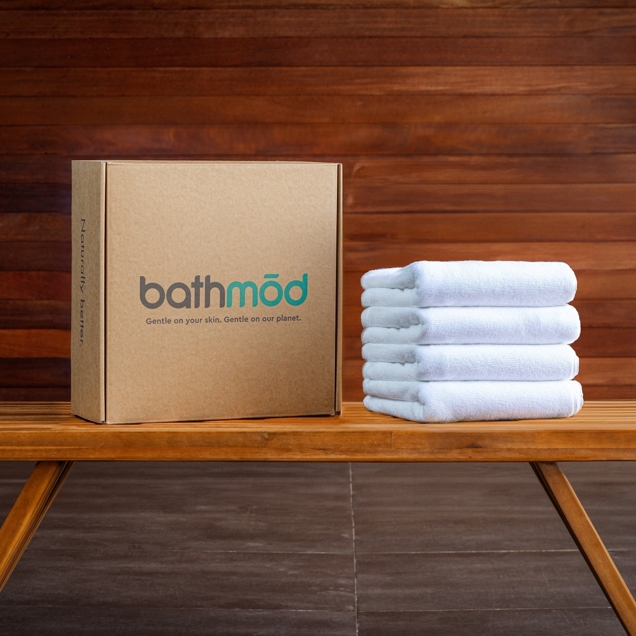 Luxury Organic Bath Mats four-pack in a cardboard box beside neatly folded white towels on wood surface